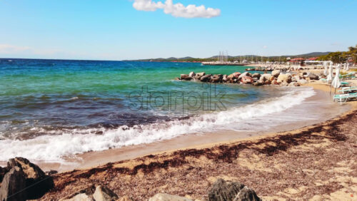 Video - Beach on Aegean sea coast with closed umbrellas and sunbeds, rocks in the water, embankment street, sea port and green hills in the distance in Nikiti, Greece. Slow motion