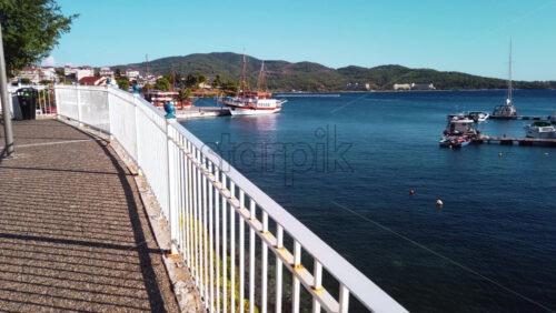 Video - View of a wide pier moored sailboat, green hill and town on the background from the pedestrian street with fence. Neos Marmaras, Greece. Slow motion
