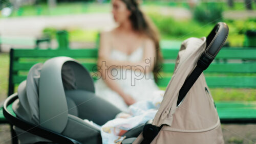 A baby resting in a stroller while a woman sits on a green bench in the background - Starpik Stock