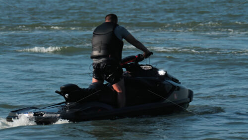 A man in a black life vest riding a black jet ski at high speed across a calm body of water - Starpik Stock