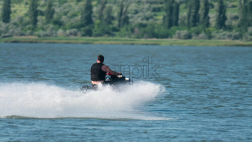 A man in a black life vest riding a black jet ski at high speed across a calm body of water - Starpik Stock