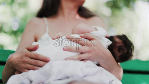 A mother siting on a bench in the park, breastfeeding her baby while enjoying a quiet, peaceful moment in nature. - Starpik Stock
