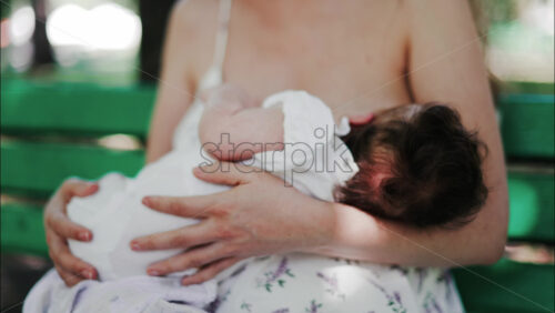 A mother siting on a bench in the park, breastfeeding her baby while enjoying a quiet, peaceful moment in nature. - Starpik Stock