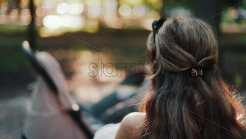 A mother with long wavy hair siting on a bench, breastfeeding her baby in a peaceful park, next to a modern baby stroller - Starpik Stock