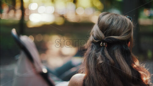 A mother with long wavy hair siting on a bench, breastfeeding her baby in a peaceful park, next to a modern baby stroller - Starpik Stock