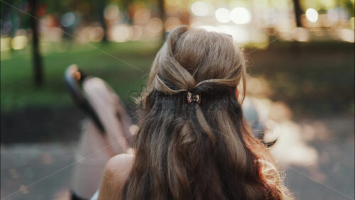 A mother with long wavy hair siting on a bench, breastfeeding her baby in a peaceful park, next to a modern baby stroller - Starpik Stock