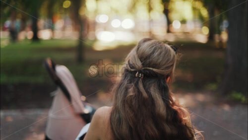 A mother with long wavy hair siting on a bench, breastfeeding her baby in a peaceful park, next to a modern baby stroller - Starpik Stock