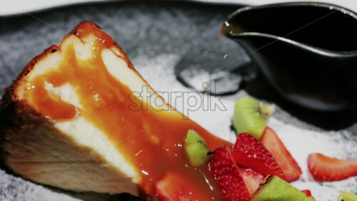 A slice of cheesecake served with caramel sauce fresh strawberries and kiwi, garnished with powdered sugar on a plate at a restaurant - Starpik Stock