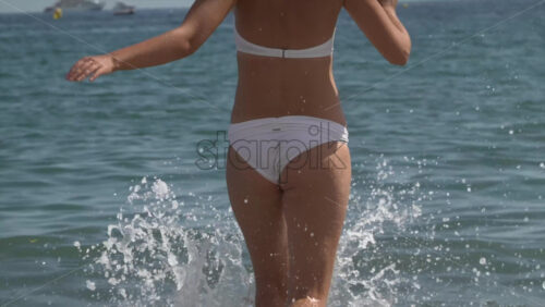 A woman in a white bikini running through knee-deep surf, sending up bright, sunlit splashes - Starpik Stock