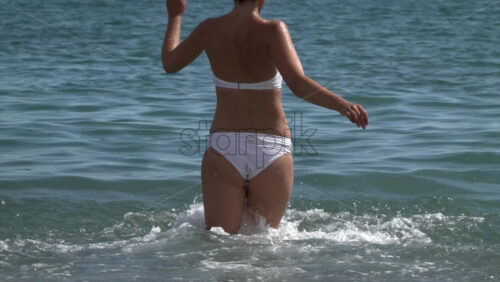A woman in a white bikini running through knee-deep surf, sending up bright, sunlit splashes - Starpik Stock