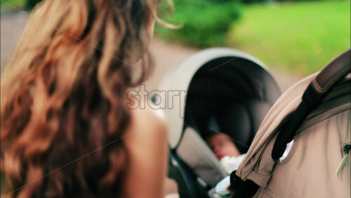 A woman with long curly hair holding a stroller while sitting on a park bench surrounded by greenery - Starpik Stock