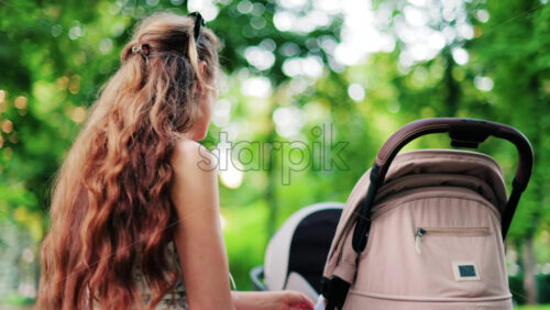 A woman with long curly hair holding a stroller while sitting on a park bench surrounded by greenery - Starpik Stock