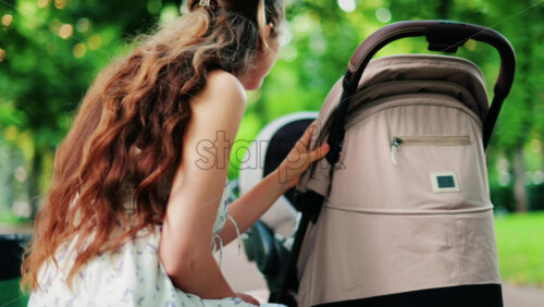 A woman with long curly hair holding a stroller while sitting on a park bench surrounded by greenery - Starpik Stock