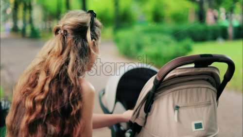 A woman with long curly hair holding a stroller while sitting on a park bench surrounded by greenery - Starpik Stock
