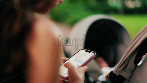 A woman with long curly hair texting on her phone while sitting on a park bench near a stroller, surrounded by greenery - Starpik Stock