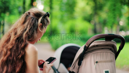 A woman with long curly hair texting on her phone while sitting on a park bench near a stroller, surrounded by greenery - Starpik Stock