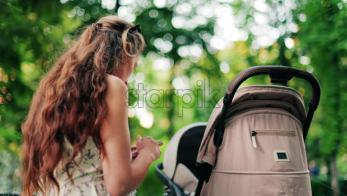 A woman with long curly hair texting on her phone while sitting on a park bench near a stroller, surrounded by greenery - Starpik Stock