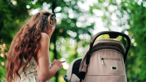 A woman with long curly hair texting on her phone while sitting on a park bench near a stroller, surrounded by greenery - Starpik Stock