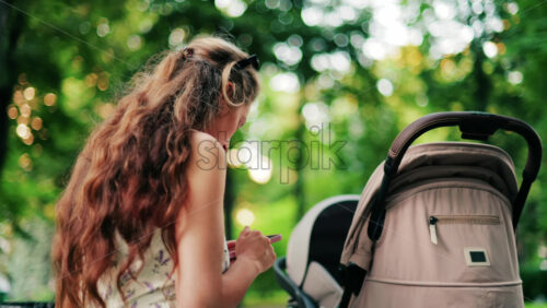 A woman with long curly hair texting on her phone while sitting on a park bench near a stroller, surrounded by greenery - Starpik Stock