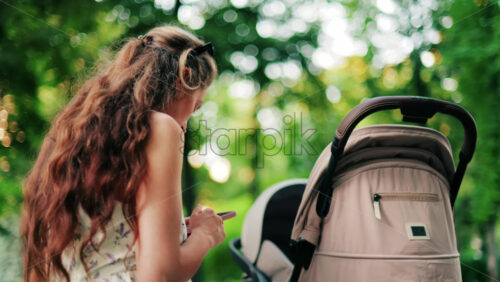 A woman with long curly hair texting on her phone while sitting on a park bench near a stroller, surrounded by greenery - Starpik Stock