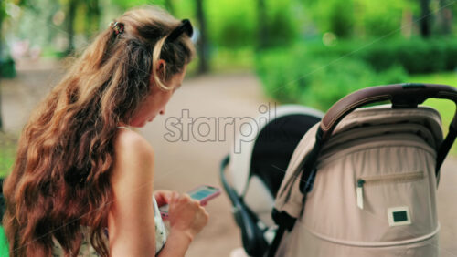 A woman with long curly hair texting on her phone while sitting on a park bench near a stroller, surrounded by greenery - Starpik Stock