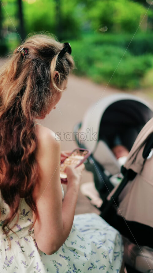 A woman with long curly hair texting on her phone while sitting on a park bench near a stroller, surrounded by greenery. Vertical - Starpik Stock