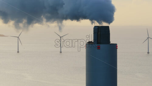 Aerial drone view of Amager Bakke power plant chimney releasing smoke, with offshore wind turbines standing in the sea behind it in Copenhagen, Denmark - Starpik Stock
