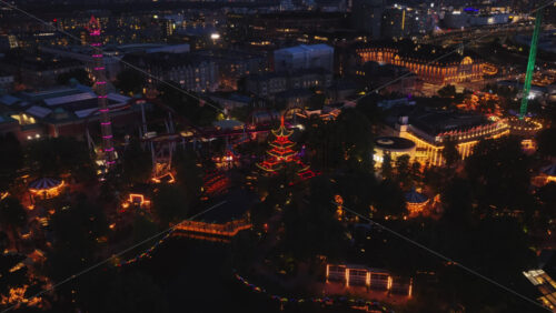 Aerial drone view of Tivoli Gardens amusement park at night, with glowing neon lights, colorful rides, and illuminated architecture in Copenhagen, Denmark - Starpik Stock