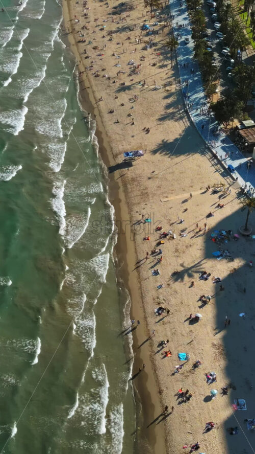 Aerial drone view of cars moving along the coastline with people relaxing on the beach in Alicante, Spain in daylight. Vertical - Starpik Stock