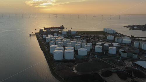 Aerial drone view of large oil storage tanks in Copenhagen Harbor, Denmark, with offshore wind turbines and the sea in the background at sunset - Starpik Stock