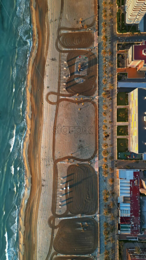 Aerial drone view of palm trees on a promenade near the beach and buildings along coastline of the city in Alicante, Spain at sunset. Vertical - Starpik Stock