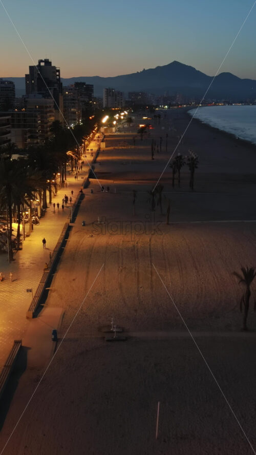 Aerial drone view of people walking on a promenade lined with palm trees near the beach in Alicante, Spain in the evening. Vertical - Starpik Stock