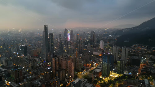 Aerial drone view of skyscrapers lighting up the skyline against the Andean foothills in Bogota, Colombia in the evening - Starpik Stock