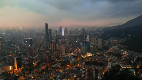 Aerial drone view of skyscrapers lighting up the skyline against the Andean foothills in Bogota, Colombia in the evening - Starpik Stock