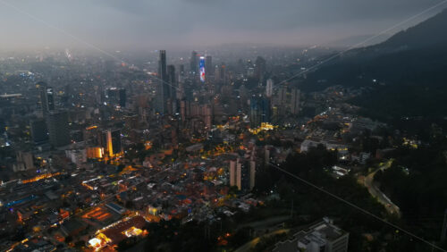 Aerial drone view of skyscrapers lighting up the skyline against the Andean foothills in Bogota, Colombia in the evening - Starpik Stock