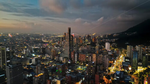 Aerial drone view of skyscrapers lighting up the skyline against the Andean foothills in Bogota, Colombia in the evening - Starpik Stock