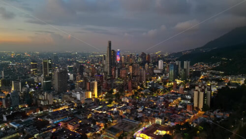 Aerial drone view of skyscrapers lighting up the skyline against the Andean foothills in Bogota, Colombia in the evening - Starpik Stock