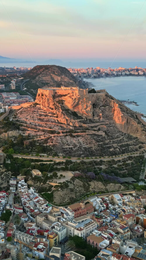 Aerial drone view of the Santa Barbara Castle surrounded by buildings on the coast of Alicante, Spain with the sea on the background. Vertical - Starpik Stock