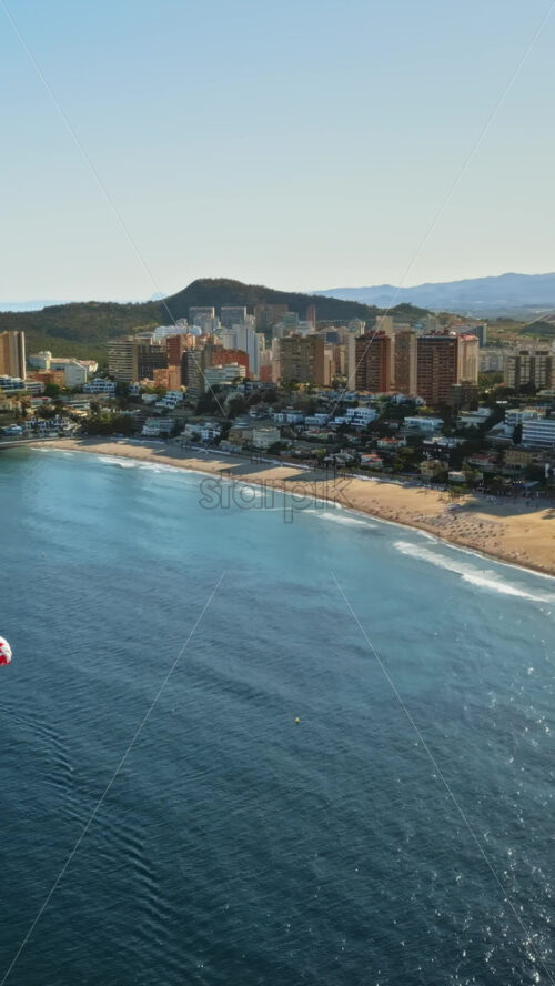 Aerial drone view of the buildings along the coastline with people parasailing in the sea in Benidorm, Spain in daylight. Vertical - Starpik Stock
