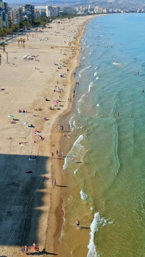 Aerial drone view of the buildings along the coastline with people relaxing on the beach in Alicante, Spain in daylight. Vertical - Starpik Stock