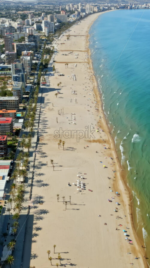 Aerial drone view of the buildings along the coastline with people relaxing on the beach in Benidorm, Spain in daylight. Vertical - Starpik Stock