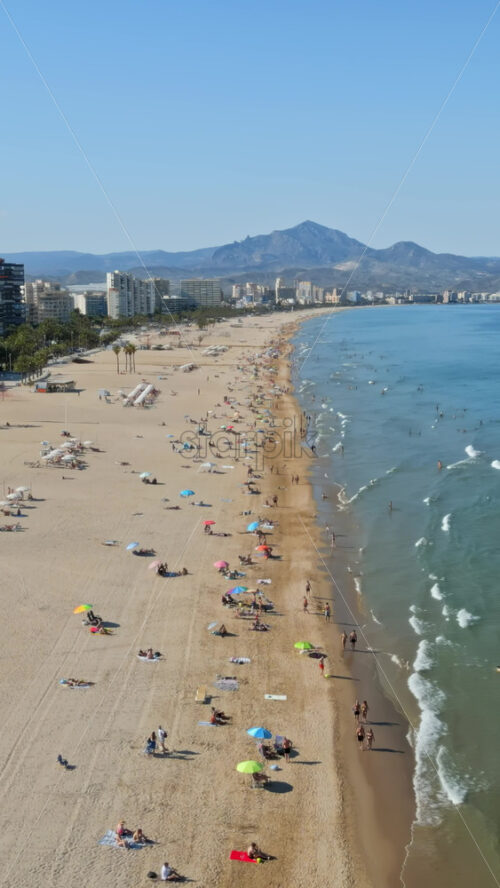 Aerial drone view of the buildings along the coastline with people relaxing on the beach in Benidorm, Spain in daylight. Vertical - Starpik Stock