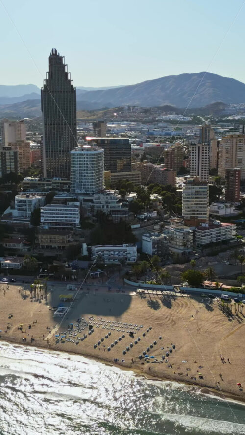 Aerial drone view of the buildings along the coastline with people relaxing on the beach in Benidorm, Spain in daylight. Vertical - Starpik Stock
