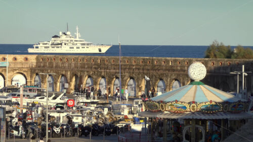 Antibes, France – July 2, 2025: Scenic coastal view featuring a luxury yacht, historic stone arches, and a colourful carousel - Starpik Stock