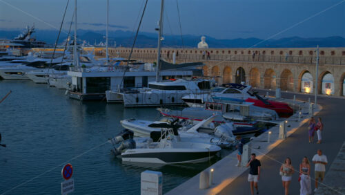 Antibes, France – June 7, 2025: People walking by multiple white boats docked in the Port Vauban with the The Nomad by Jaume Plensa sculpture on the background in the evening - Starpik Stock