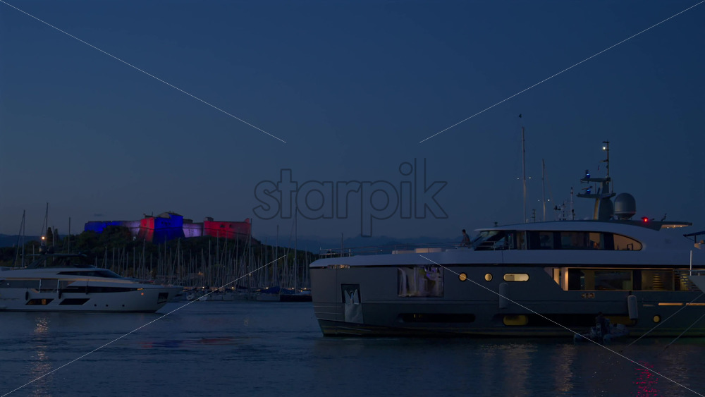 Antibes, France – June 8, 2025: Boats moving through Port Vauban with Fort Carre illuminated on the background in the evening - Starpik Stock