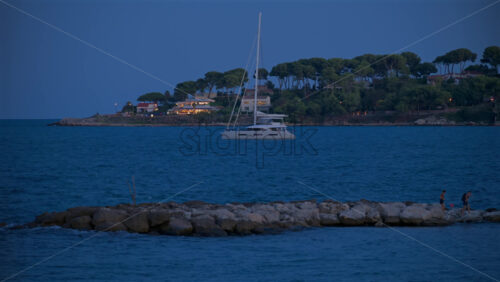 Antibes, France – June 8, 2025: People walking on a rocky breakwater with a white boat floating on the Mediterranean sea on the background in the evening - Starpik Stock