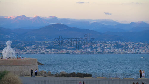 Antibes, France – June 8, 2025: People walking on the coast of the city with The Nomad by Jaume Plensa sculpture on the background - Starpik Stock