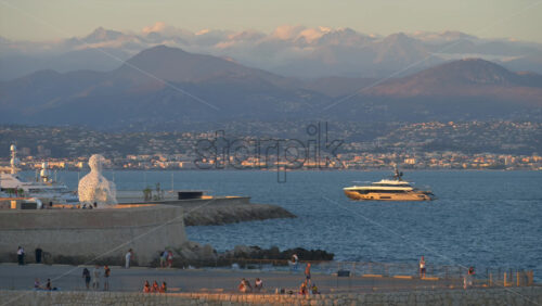 Antibes, France – June 8, 2025: People walking on the coast of the city with The Nomad by Jaume Plensa sculpture on the background - Starpik Stock