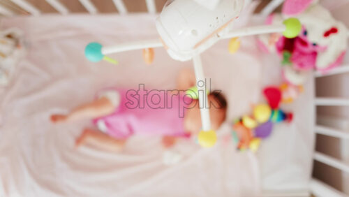 Baby lying on a bed in a pink outfit, looking up at a colourful hanging mobile toy - Starpik Stock